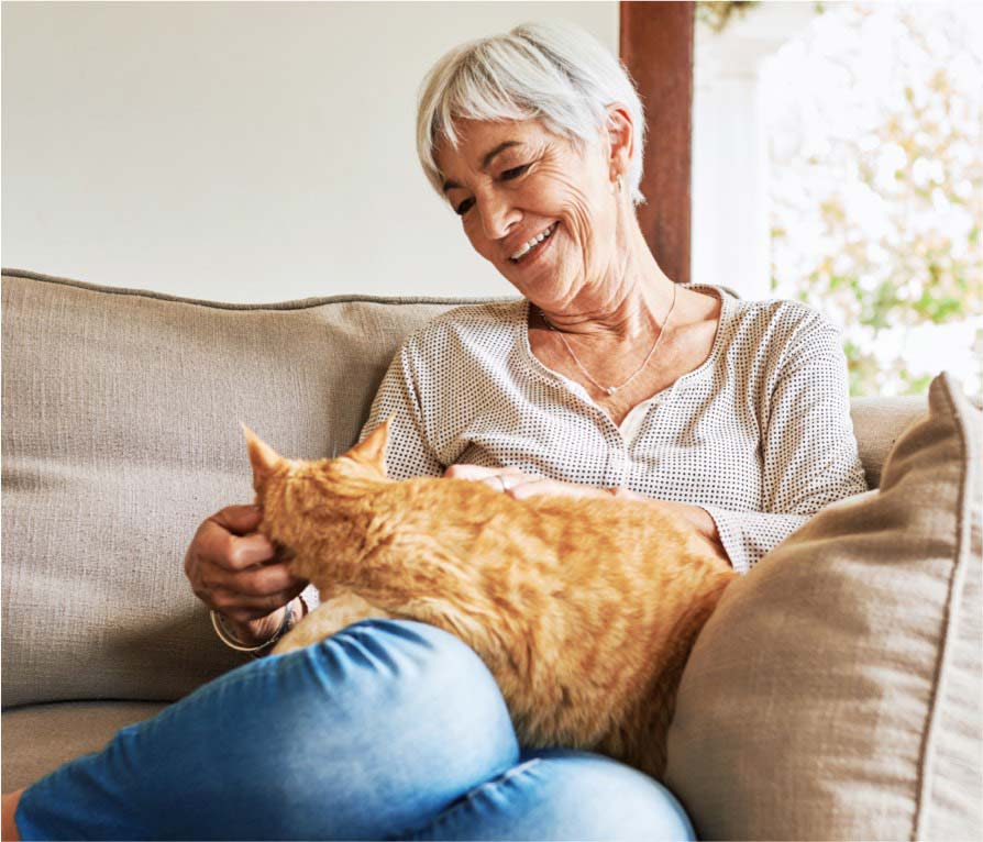 Elderly woman smiling while petting an orange cat on her lap, sitting on a beige couch.
