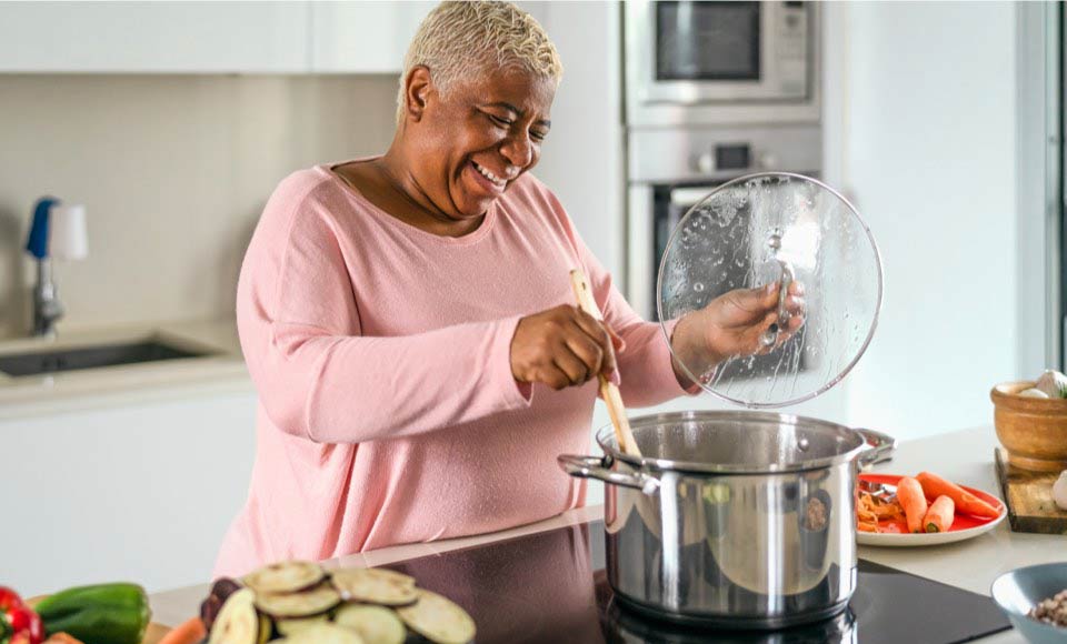 A senior woman is smiling while cooking with a large pot in a modern kitchen.