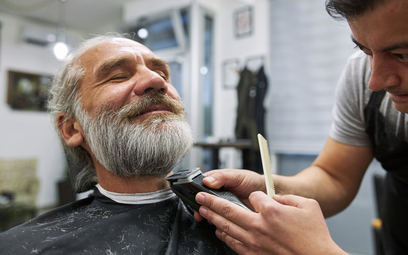 Barber trims an older mans beard with an electric clipper in modern, well-lit salon