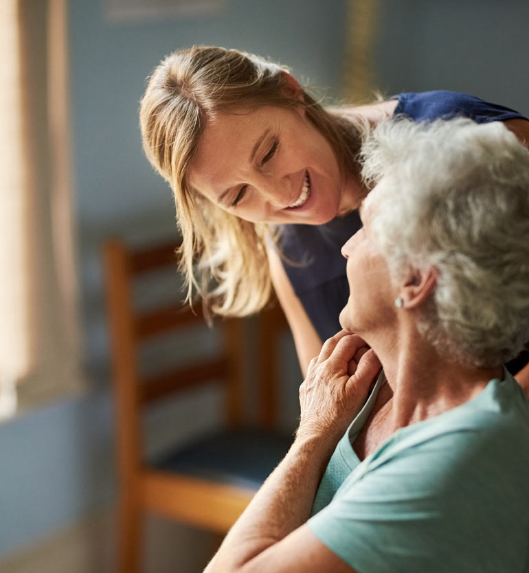 Caregiver smiling warmly at a senior woman in a cozy room during a caregiving session.