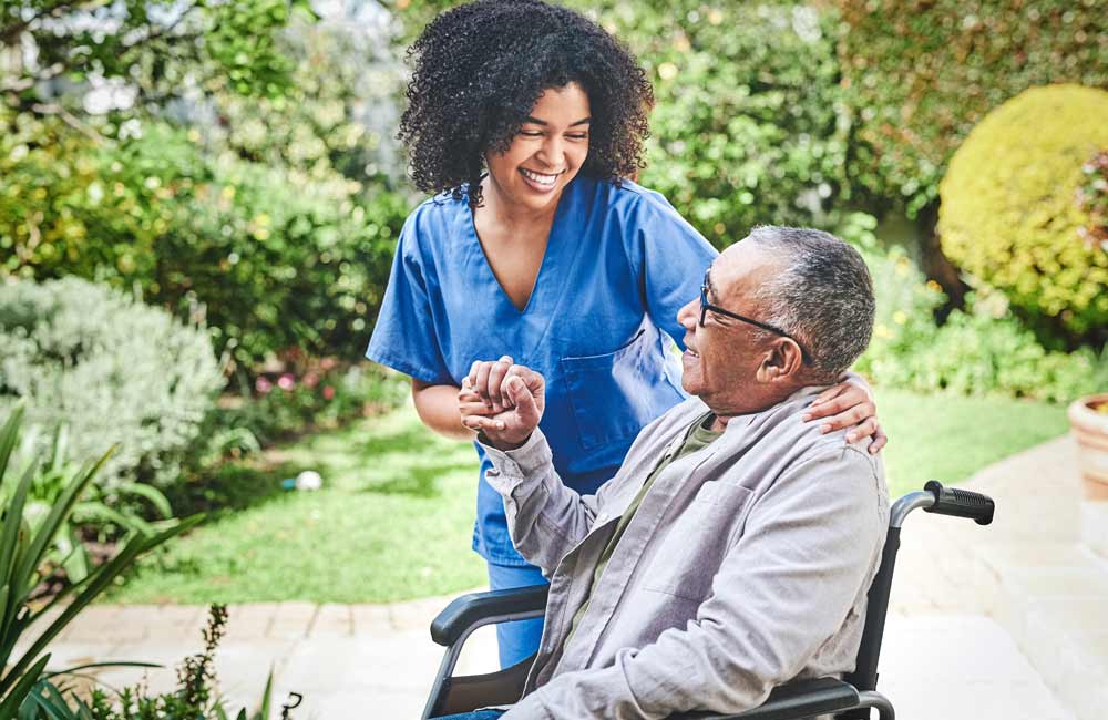 Caregiver in blue scrubs smiling at a senior man in a wheelchair in an outdoor garden