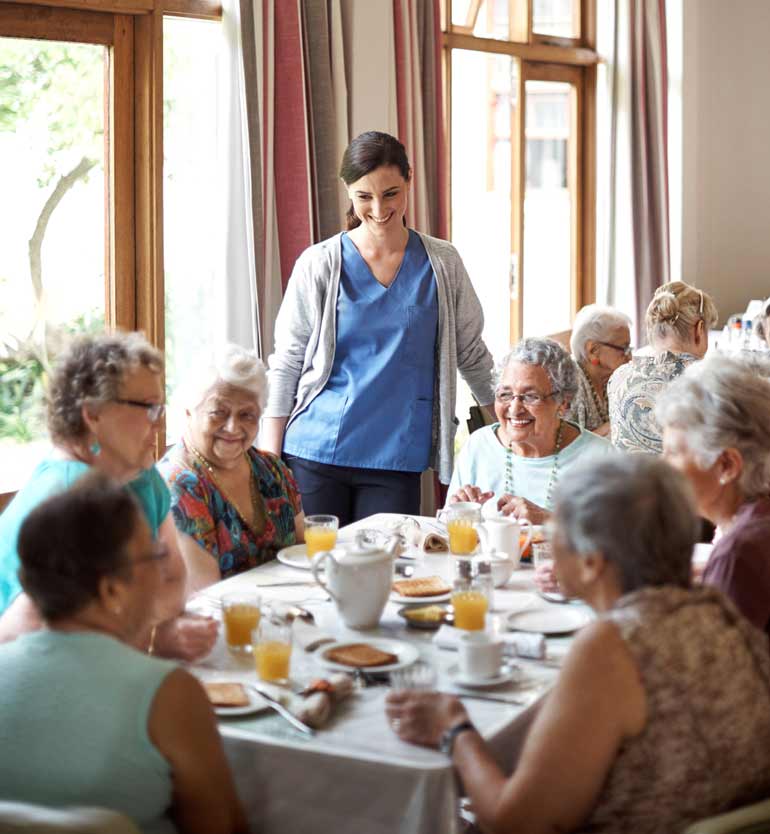 A group of senior women dining together, assisted by a caregiver in a cozy dining room.