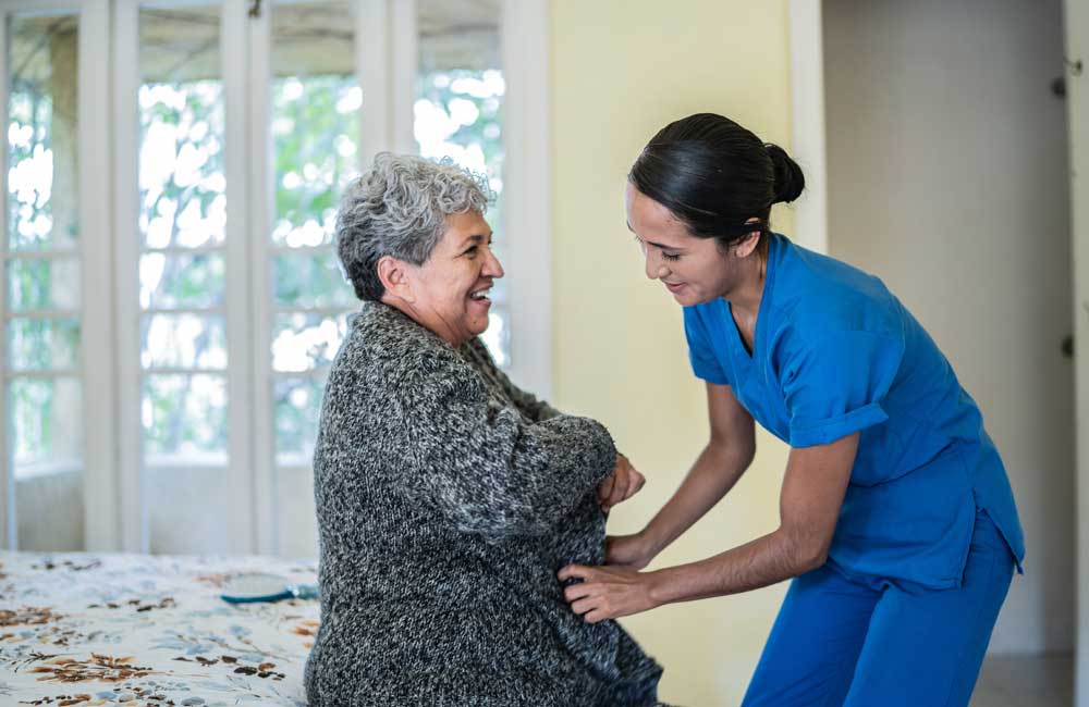 A smiling elderly person receives assistance from a caregiver in blue scrubs inside a bright room.