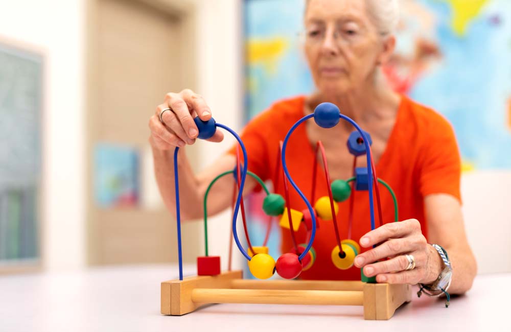 Elderly woman in orange shirt participates in memory care activity with colorful bead toy.