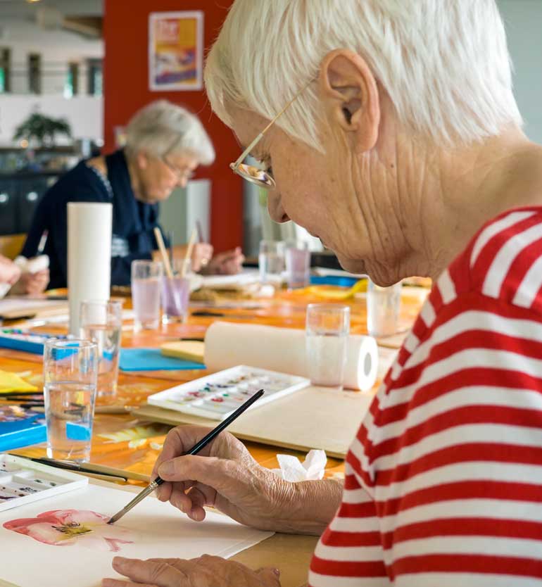 Elderly woman painting a pink watercolor flower in art class