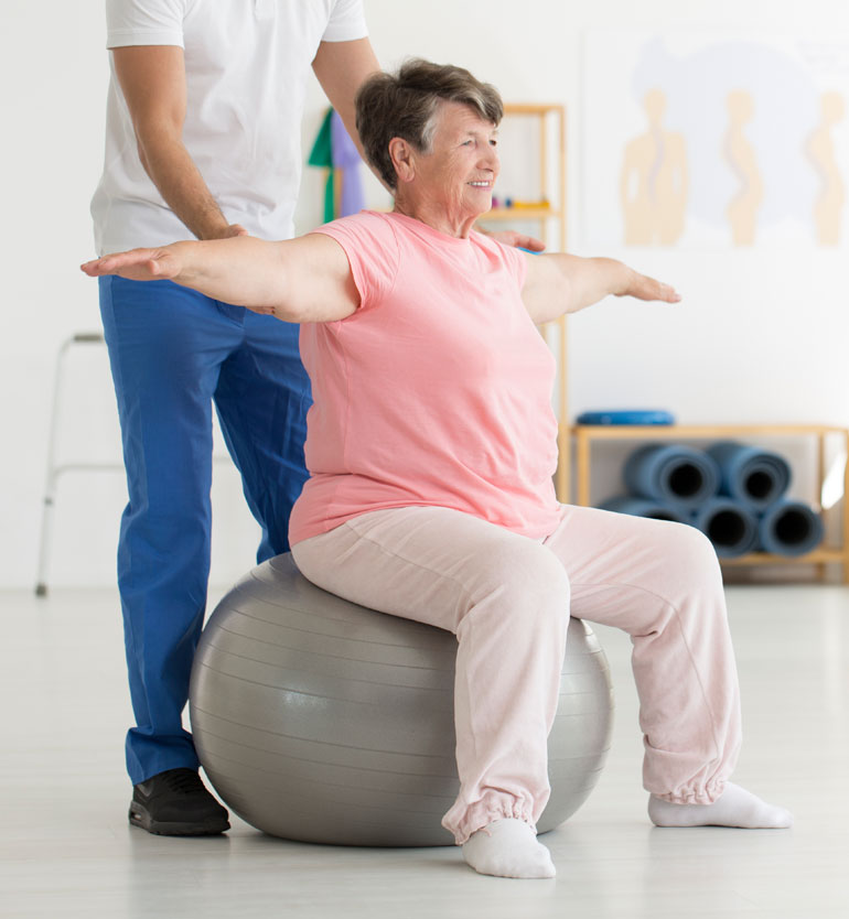 Elderly woman in pink doing physical therapy exercises on a stability ball with therapist support.