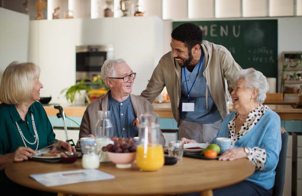Senior care provider engaging with elderly individuals at a table during a meal time.