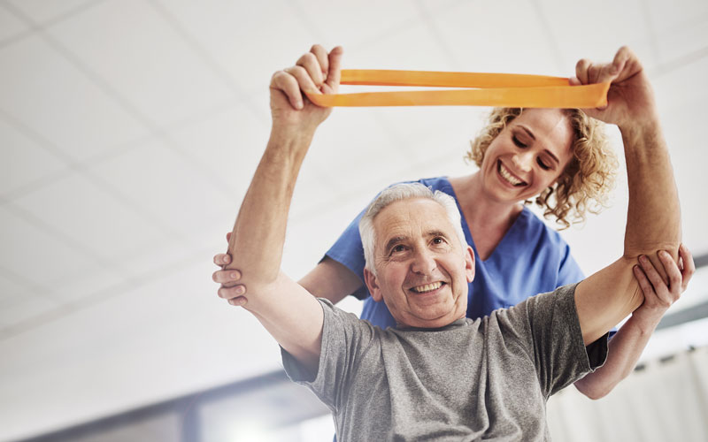Senior man exercising with resistance band assisted by a smiling caregiver in blue scrubs.