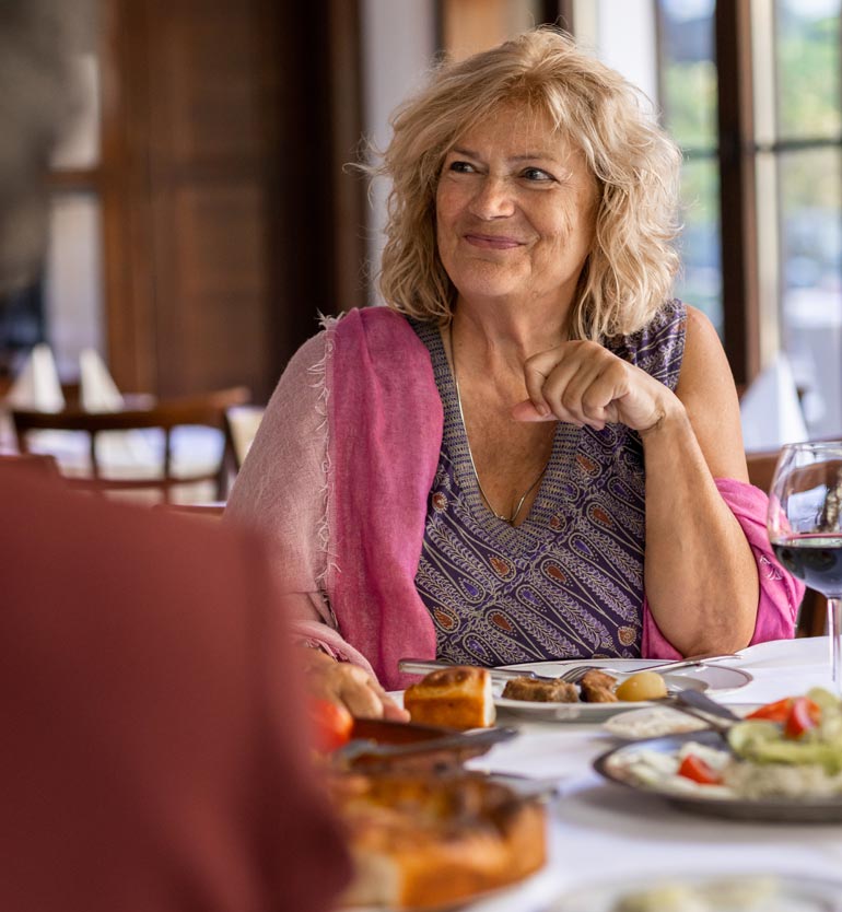 Senior woman enjoying a meal with friends at a restaurant, smiling and wearing a pink shawl.