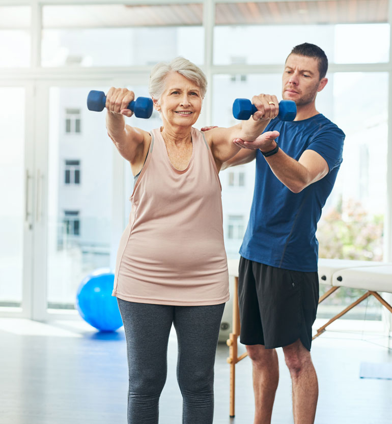 Senior woman lifting dumbbells with a trainers assistance in a bright exercise room.