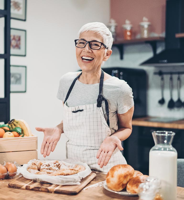 Smiling senior woman wearing apron and presenting freshly baked goods in a modern kitchen