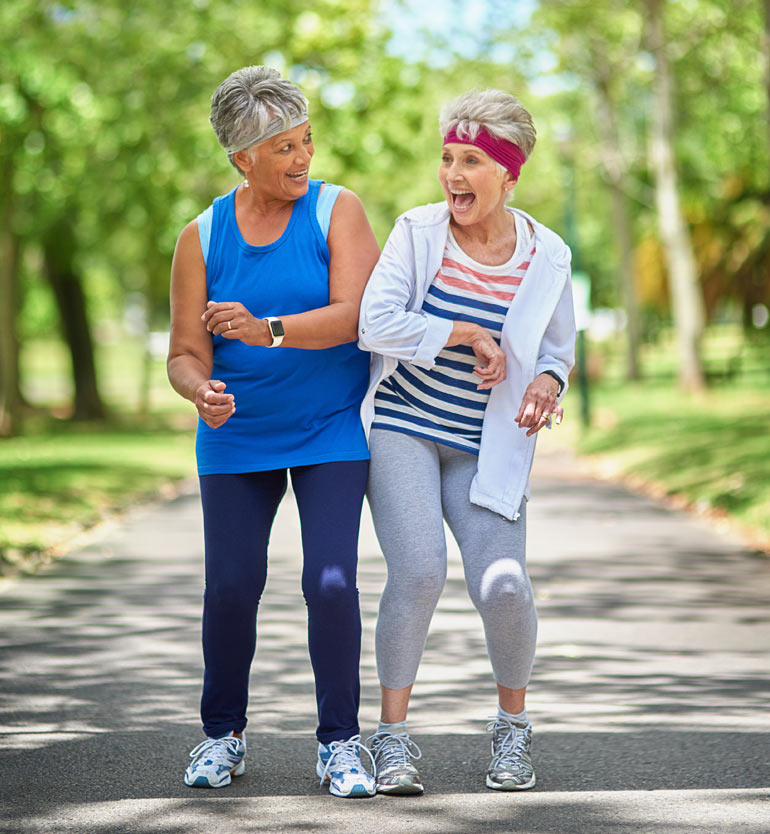 Two senior women laughing and exercising together in a sunny park.