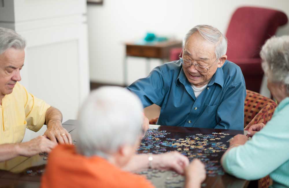 Four senior individuals are enjoying putting together a jigsaw puzzle on a table at home.