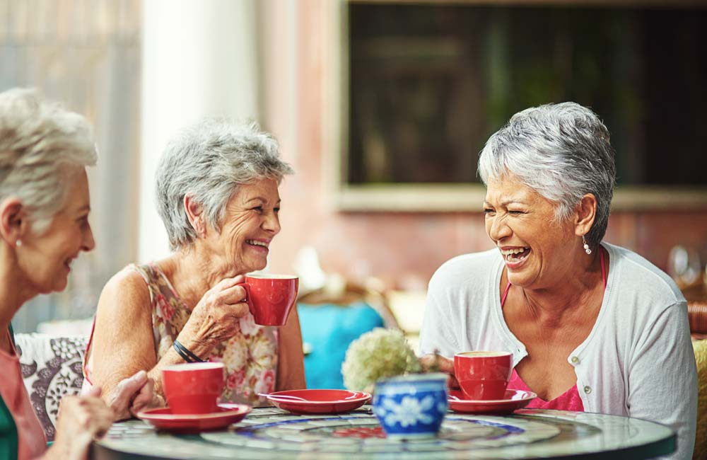 Three senior women laughing and enjoying coffee together at an outdoor table.