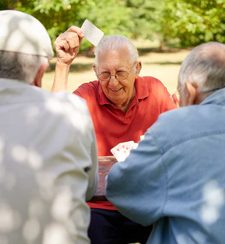 Three seniors enjoy a lively game of cards outdoors on a sunny day at a wooden table.