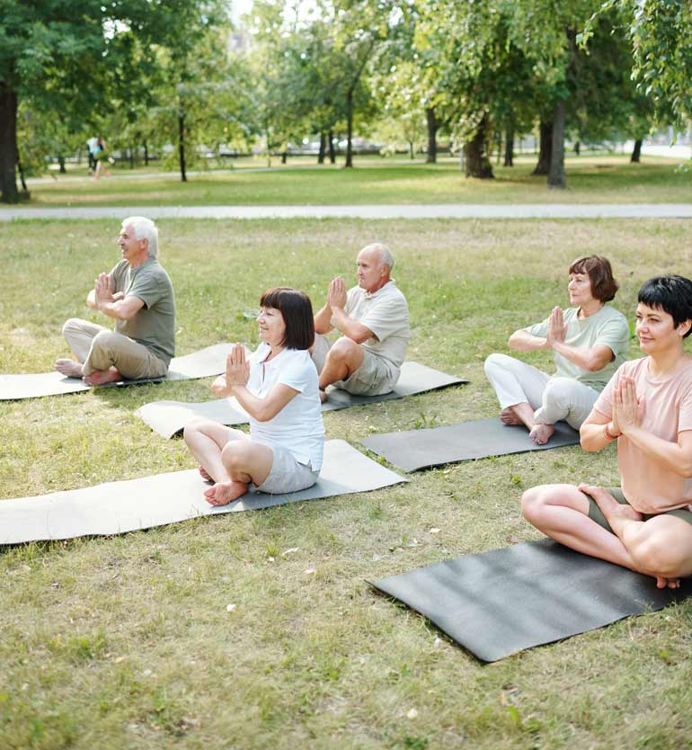 Group of seniors practicing yoga outdoors, sitting on mats with hands in prayer position