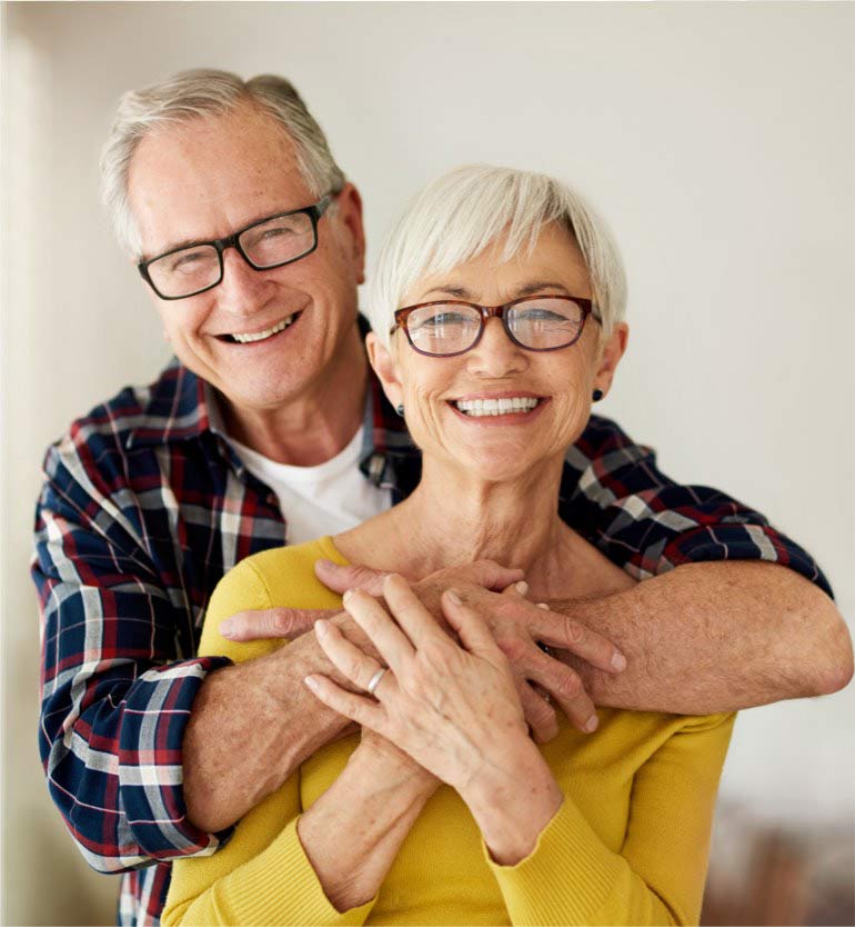 Smiling senior couple embracing indoors, wearing glasses and casual clothing.